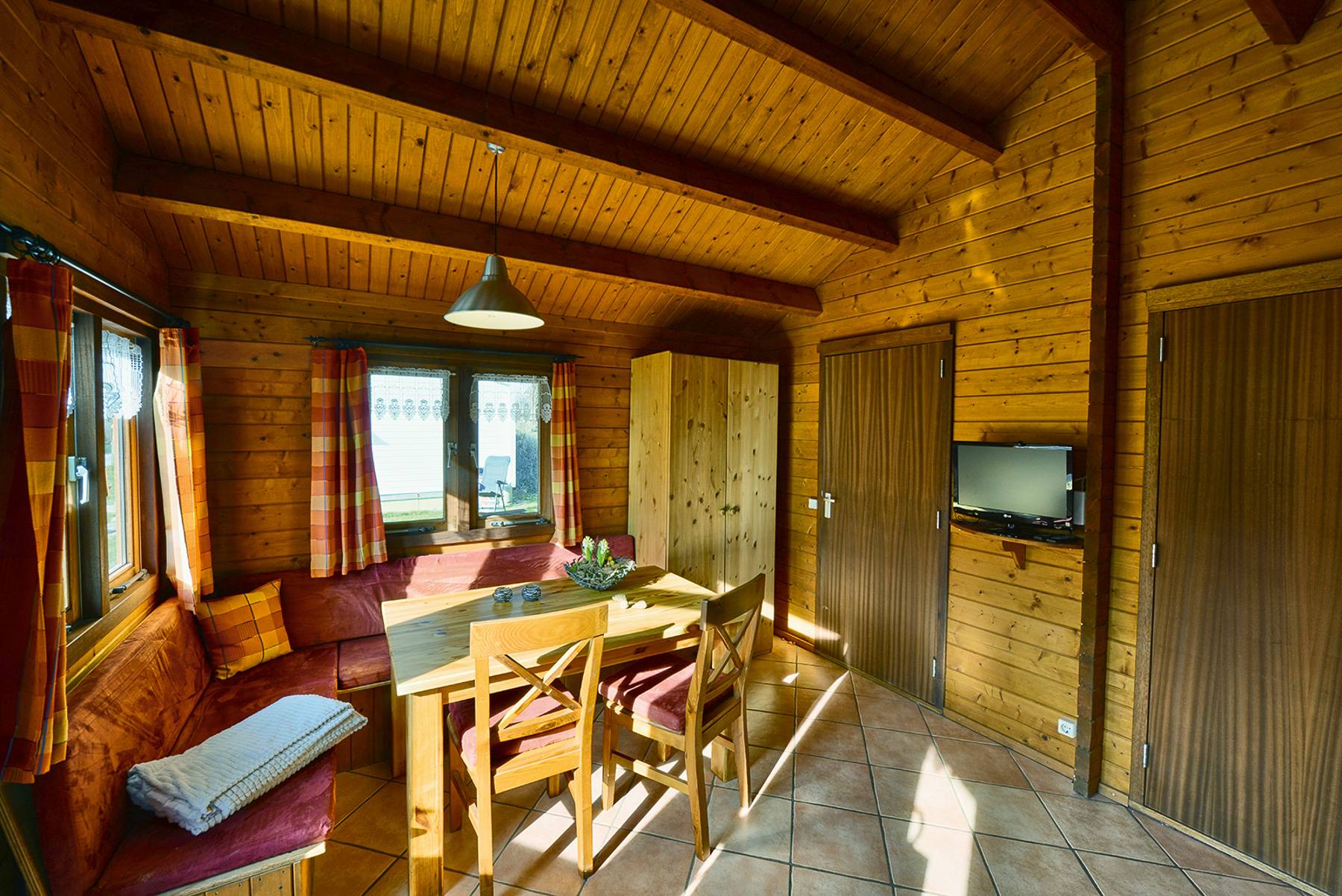 Gemütlicher Innenraum einer Wanderhütte mit viel Holz, warmem Licht und großen sonnigen Fenstern.